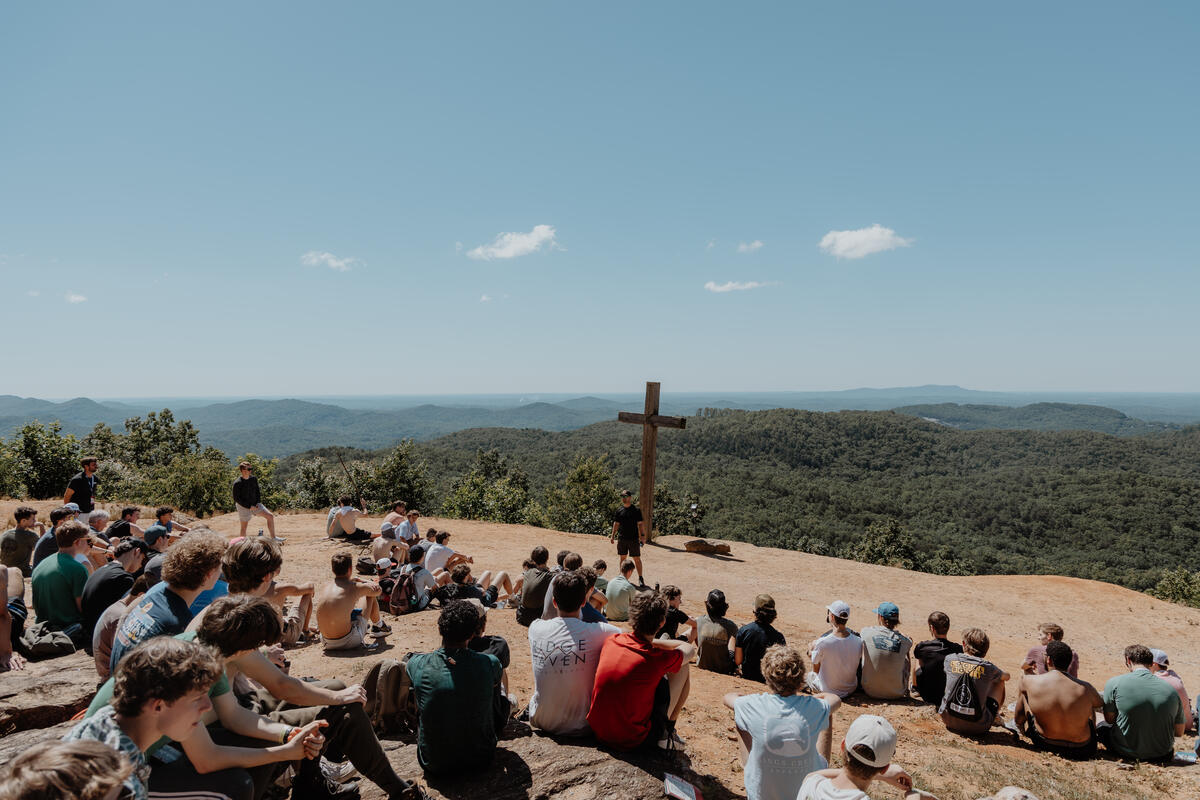 Students sitting on the rock overlook around a cross, wooded hills behind.